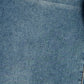 A close-up of blue denim fabric texture with subtle fading and a small portion of contrasting stitching visible at the lower right edge.