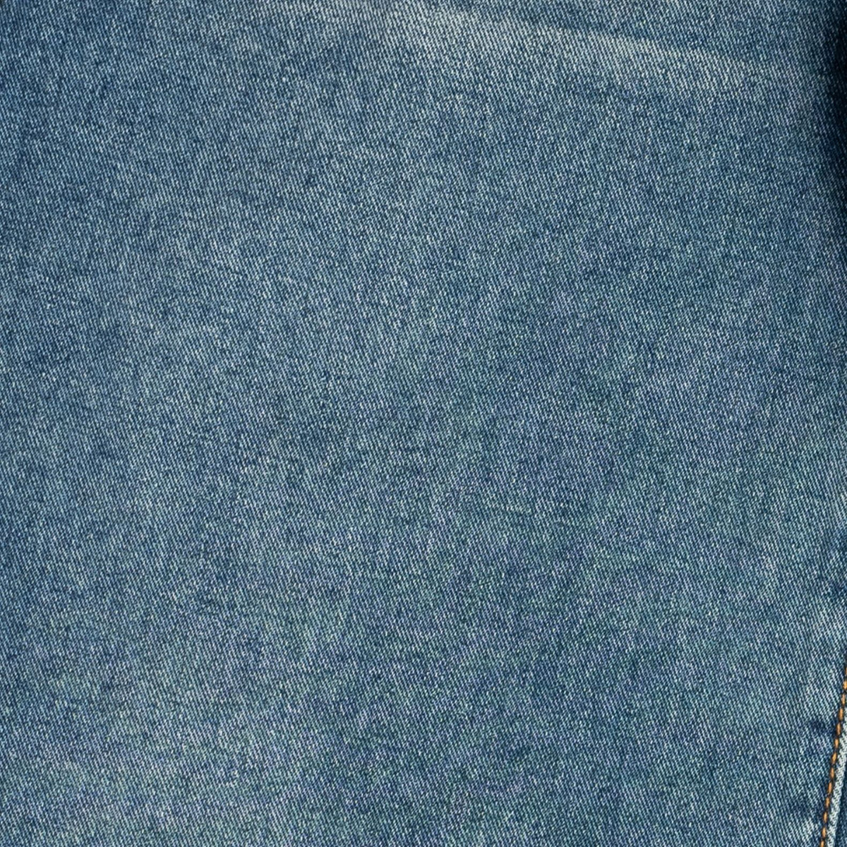 A close-up of blue denim fabric texture with subtle fading and a small portion of contrasting stitching visible at the lower right edge.
