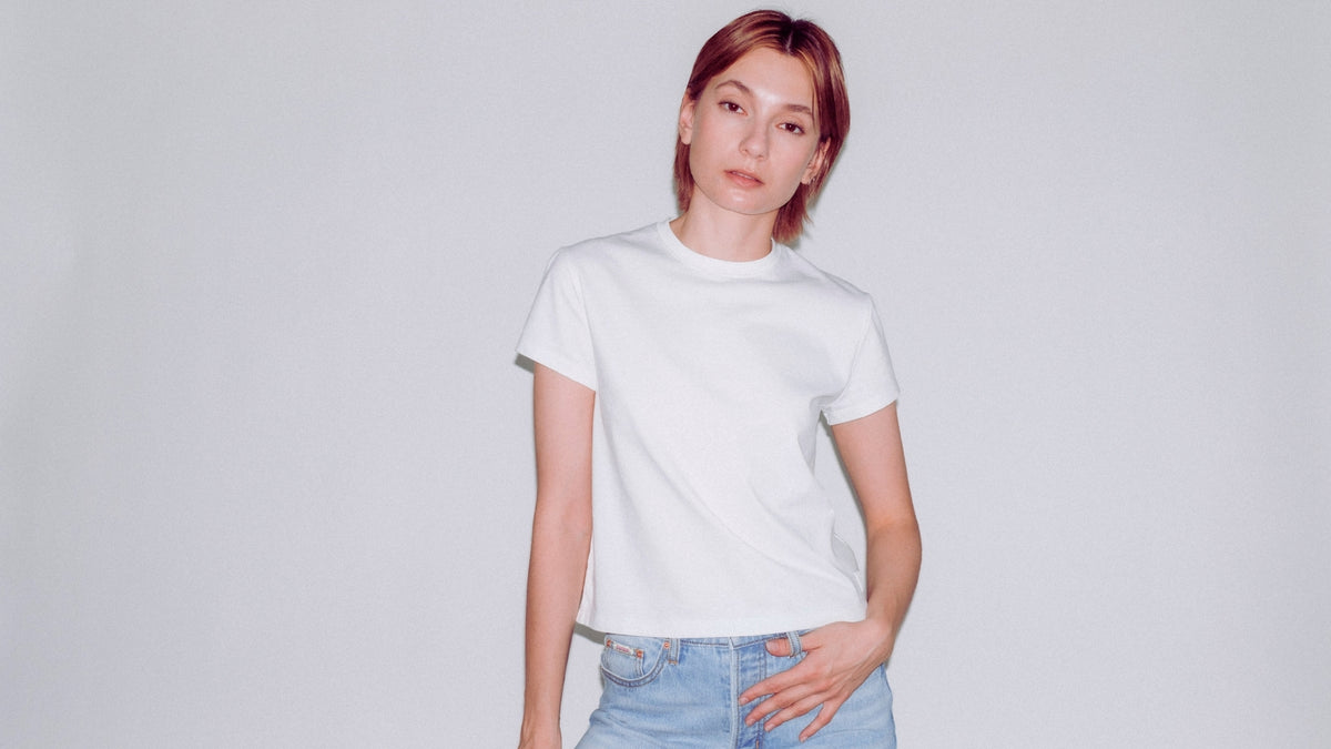 A model with short reddish-brown hair wearing a plain white short-sleeve crewneck t-shirt and light blue Bubblegum jeans, standing against a light gray studio background.