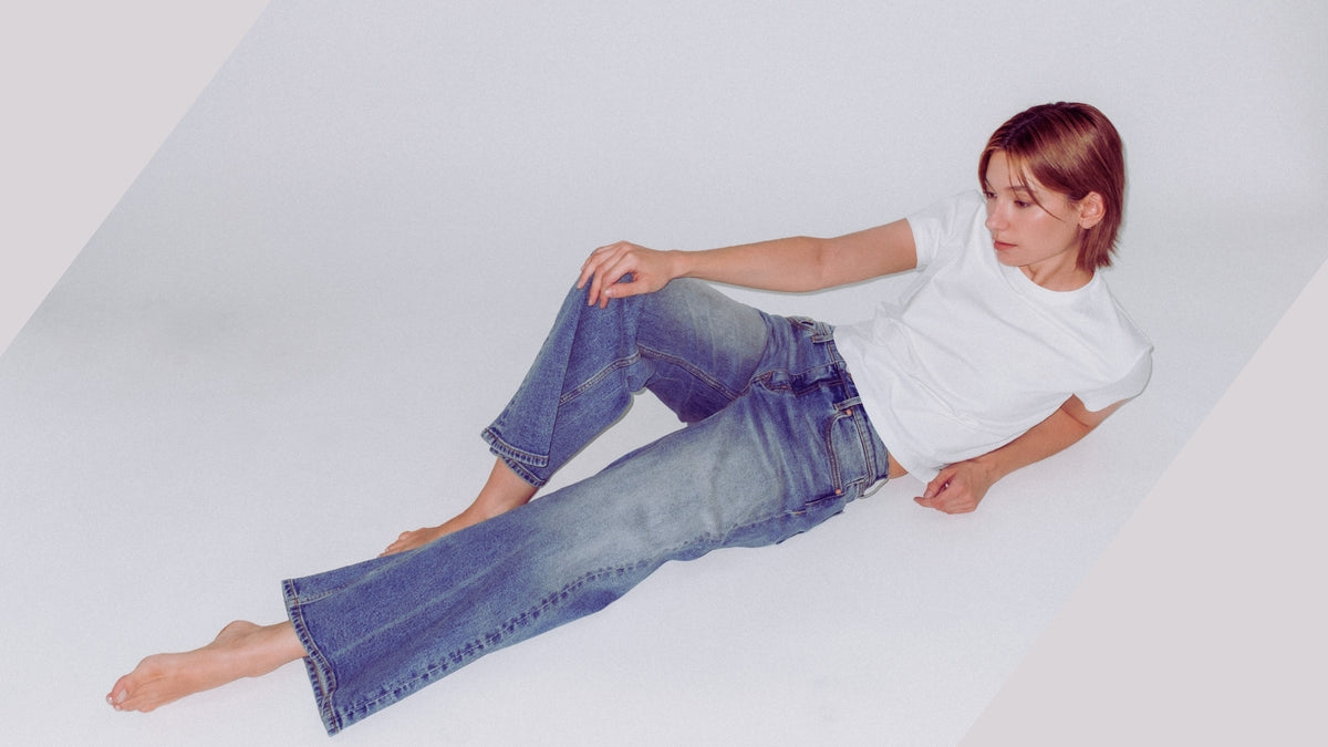 A woman with short brown hair lies on her side on a white studio floor, wearing a white short-sleeve T-shirt and blue wide-leg Bubblegum jeans.