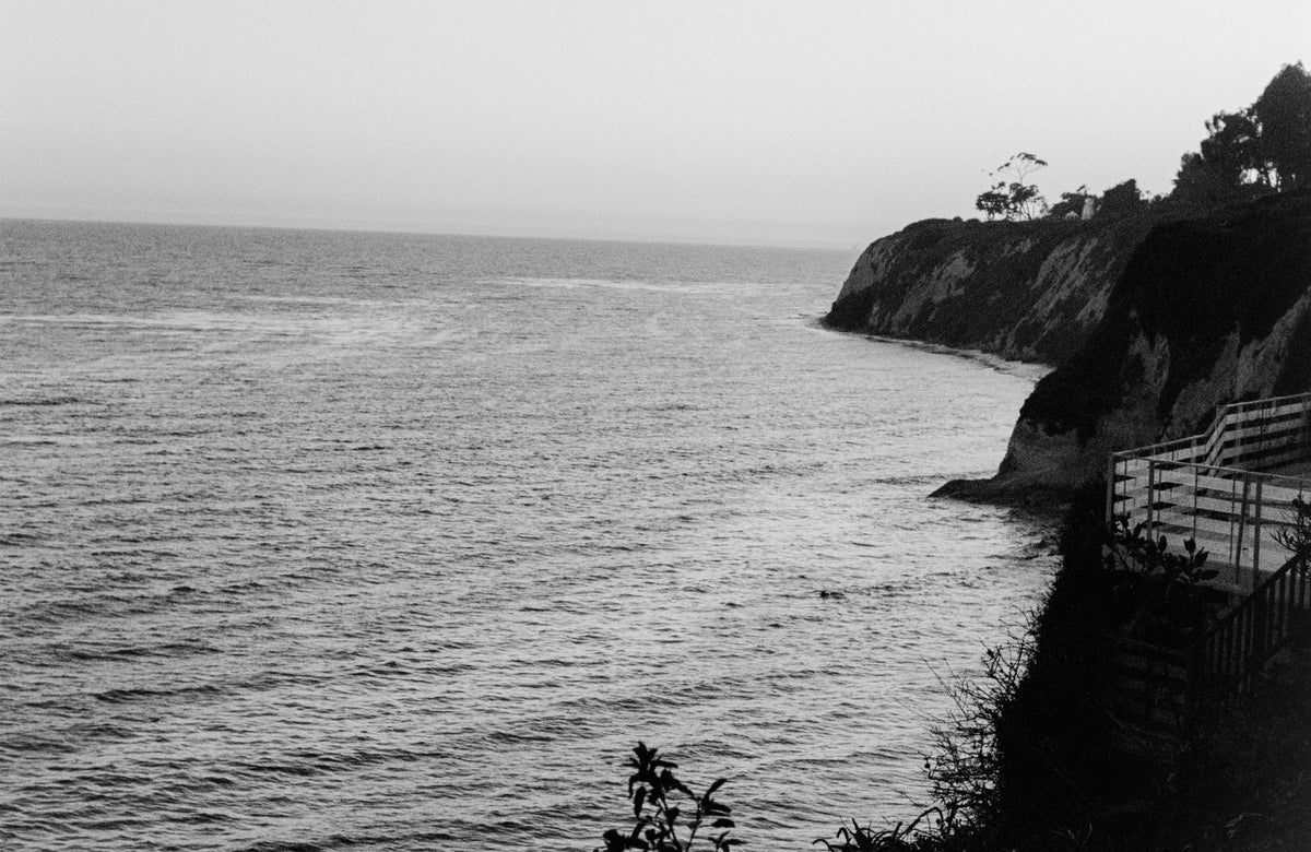 Black and white photo of a coastal scene with a cliff and ocean. Here since '96. Every fade has a story. Bubblegum USA.